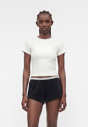 Λεπτός άνθρωπος με σκούρο δέρμα και πλεξούδες, φορώντας εφαρμοστό λευκό cropped t-shirt και μαύρα σορτς με ζώνη "Juicy Couture".