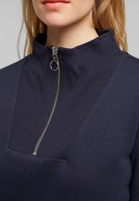 Mörkblå zip-up sweatshirt med hög krage, ribbad textur och en silverdragkedja vid halsringningen.