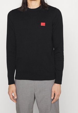 Henkilö, jolla on musta pitkähihainen pyöreäkauluksinen neulepaita, jossa on punainen "HUGO"-logopatch rinnassa, ja vaaleanharmaat housut valkoista taustaa vasten.