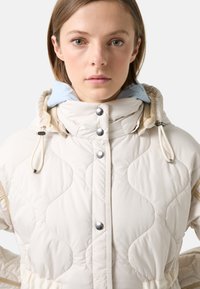 Quiltet hvid jakke med hætte, prydet med sølv trykknapper og et lysblåt foer. Tekstureret stof med justerbare snore.