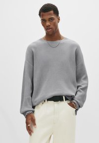 Junger Mann in einem lockeren grauen Strickpullover, cremefarbenen Hosen und silberner Schmuck, der mit einer Hand in der Tasche vor einem weißen Hintergrund steht.