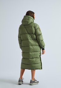 Garšs olīvu zaļš pūkainais mētelis ar kapuci, quiltēta dizaina, sānu šķēlumi un uzlīme kreisajā plecā. Valkāts ar pelēkiem kedām.