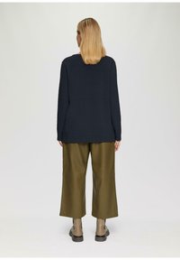 Blonde vrouw staat met haar rug naar ons toe, gekleed in een zwart shirt met lange mouwen, brede olijfgroene broek en beige enkellaarsjes op een grijze achtergrond.