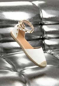 Weiße Espadrille-Flatsandale mit Knöchelriemen und goldenen Nieten, vor einem glänzenden silbernen gepolsterten Hintergrund präsentiert.