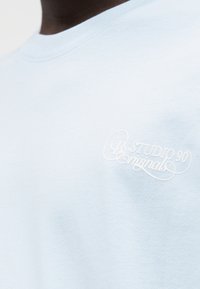 Φωτεινή γαλάζια βαμβακερή μπλούζα με στρογγυλή λαιμόκοψη και κεντημένο λογότυπο "Studio 90 Originals" σε λευκό στο στήθος. Μαλακή υφή.