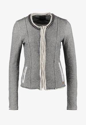 Frau trägt graue Jacke über hellgrauem Hemd, marineblaue Hose, schwarze Stiefeletten, hält eine dunkle Clutch-Tasche.