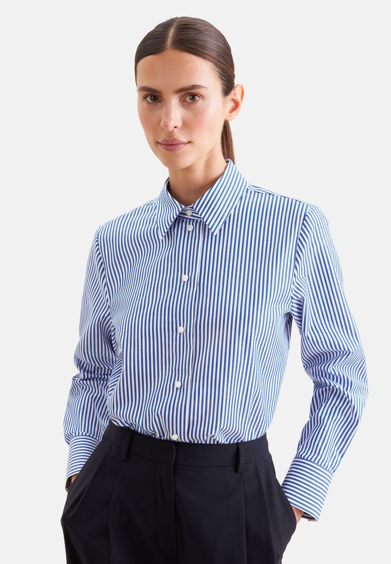 Femme aux cheveux foncés attachés en queue de cheval portant une chemise à boutons rayée verticalement bleue et blanche, rentrée dans un pantalon noir taille haute.
