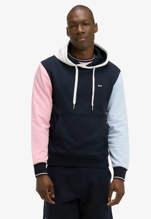 Homme portant un sweat à capuche bleu marine avec une capuche blanche, une manche gauche rose, et une manche droite bleu clair, debout devant un fond uni.