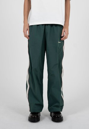 Persona con pantalones sueltos verde oscuro con franjas laterales blancas y rojas, camiseta blanca y zapatos negros gruesos, de pie contra un fondo gris.