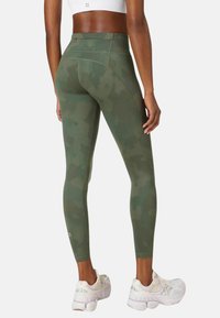 Groene camouflageleggings van rekbare stof, met een hoge tailleband en een gladde textuur. Draagt witte sportschoenen.