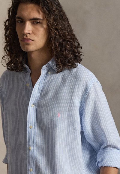 Jonge man met lang, krullend haar die een lichtblauw, witgestreept overhemd met opgerolde mouwen draagt tegen een effen achtergrond.