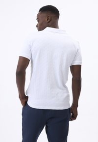 Camisa polo blanca con un patrón texturizado, mangas cortas y un cuello clásico. Combinada con pantalones azul oscuro, vista desde atrás.