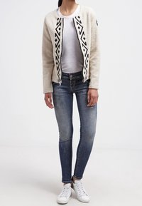 Miękki kremowy kardigan z czarnym geometrycznym wzorem, zapinany na zamek, zestawiony z niebieskimi dżinsami i białymi trampkami. Gładka tekstura, luźny krój.