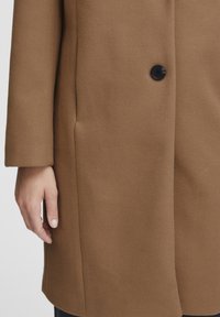 Cappotto marrone realizzato in tessuto morbido, con un solo bottone nero, taglio dritto e tasche laterali con una texture liscia.