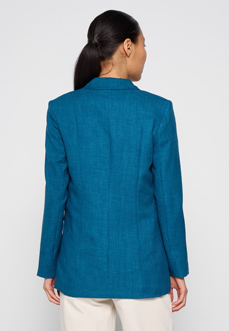 Blazer teal avec une finition texturée, présentant un col classique et une coupe ajustée, des détails de coutures visibles et des manches longues.