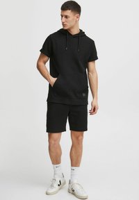Sort kortærmet hoodie med tekstureret overflade og frontlomme, parret med matchende sorte shorts. Modellen bærer hvide sneakers og sokker.