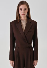 Bruine getailleerde blazer met een enkele knoop en een borstzak, gecombineerd met een geribd beige top. Glad materiaal met een gestructureerde pasvorm.