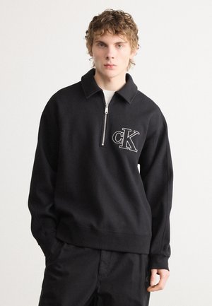 Pulover scurt din bumbac negru cu fermoar la gât, având un guler și un logo alb contrastant "CK" pe pieptul stâng, precum și manșete cu dungi.