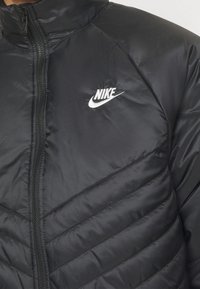 Svart vadderad jacka av slät tyg med hög krage. Har ett diagonalt quiltat mönster och en vit Nike-logotyp på bröstet.