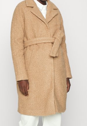 Persona con abrigo camel texturizado hasta la rodilla, con cuello ancho, cintura ceñida con cinturón, mangas largas, sobre camisa blanca y pantalones.