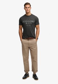 Dunkelgraues Baumwoll-T-Shirt mit "Superdry & Co"-Druck, braune Chinos und dunkle Schuhe. Das T-Shirt hat kurze Ärmel und einen Rundhalsausschnitt.