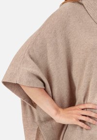 Poncho tricoté beige avec un col côtelé, des manches surdimensionnées et des fentes sur les côtés. Texture douce, tombe librement et offre une coupe décontractée.