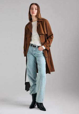 Femme portant un trench marron, un top blanc froncé, un jean large bleu clair, des bottines noires et tenant un petit sac imprimé léopard.