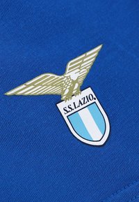 Tissu bleu avec un emblème d'aigle doré au-dessus d'un bouclier comportant deux bandes verticales bleu clair et le texte "S.S. LAZIO" en dessous.
