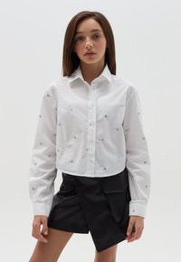 Camisa blanca de botones con bordado de corazón plateado, diseño corto y mangas largas; combinada con pantalones cortos negros tipo wrap que tienen un bolsillo lateral.