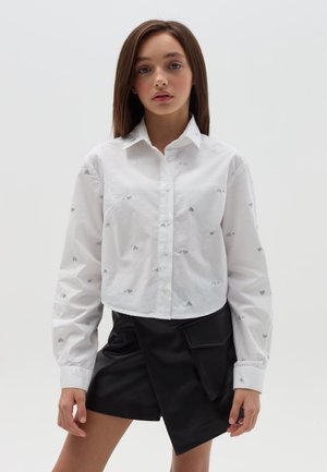 Weißes Hemd mit Knöpfen, verziert mit silberner Herzstickerei, im cropped Design und mit langen Ärmeln; kombiniert mit schwarzen Wickelhosen, die eine Seitentasche haben.