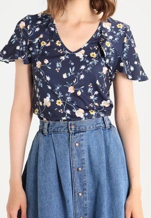 Frau trägt eine dunkelblaue, florale Kurzarmbluse mit Rüschenärmeln und einen hoch taillierten, blauen Jeansrock mit Knopfleiste vorne.