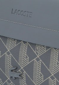 Borsa grigia dalla texture con motivo geometrico in rilievo, impreziosita da un logo Lacoste in rilievo e da un emblema a forma di coccodrillo.