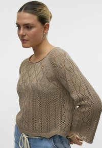 Maglione beige lavorato a maglia con un motivo a rombi aperti, maniche lunghe e scollatura rotonda. Indossato con jeans azzurri e orlo con cordoncino.
