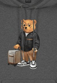 La grafica presenta un orsetto stilizzato che indossa occhiali, una maglietta nera, pantaloni beige e sneakers, mentre tiene una valigia decorata, su un hoodie grigio.