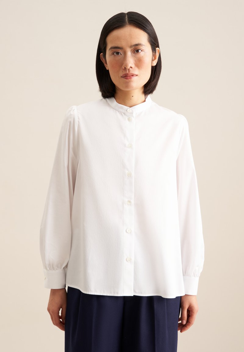 Blusa bianca con bottoni, colletto alla cinese, maniche lunghe e lievi dettagli a palloncino sulle spalle. Tessuto liscio e orlo leggermente arcuato.