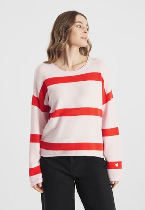 Kobieta w różowym swetrze z szerokimi czerwonymi poziomymi paskami oraz małym serduszkiem na rękawie, w czarnych spodniach, patrząca bokiem.