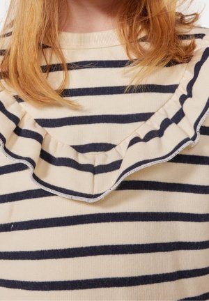 Mädchen in einem cremefarbenen Pullover mit schwarzen horizontalen Streifen und einem Rüschendetail an der Vorderseite, mit schulterlangem rötlichem Haar.
