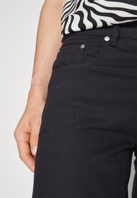 Vaqueros de mezclilla en un azul marino oscuro con una textura lisa, que presentan bolsillos frontales, herrajes metálicos y un primer plano de una mano sosteniendo la cintura.