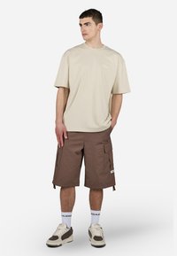 Beige bomull t-skjorte med korte ermer og hvit logo, parret med brune cargo-shorts med flere lommer og snor i bunnen.