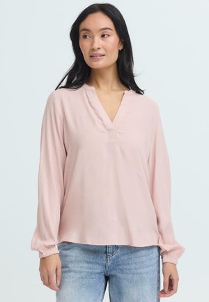 Mujer con cabello largo y oscuro que lleva una blusa de manga larga de color rosa claro con cuello en V y jeans azules, mirando de lado.