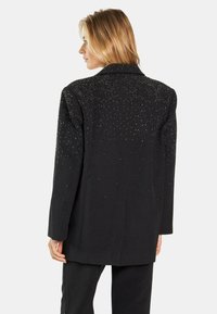 Blazer negro con collar, adornado con strass dispersos en la parte posterior, con un corte a medida y textura suave.