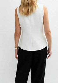 Nainen, jolla on vaaleat hiukset, pukeutunut olkaimettomaan valkoiseen toppiin ja mustiin housuihin, kuvattuna selkäpuolelta vaaleaa taustaa vasten.