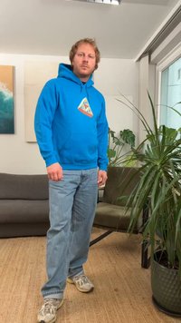 Sweatshirt azul com um gráfico triangular em laranja e amarelo, combinada com jeans azul claro e sapatilhas cinzentas; ambiente interior com plantas.