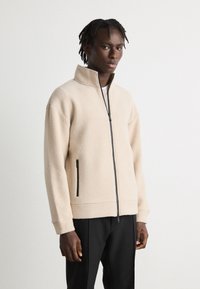 Beige fleece zip-up jacket with high collar, black zipper, and side pockets; relaxed fit with ribbed cuffs. Worn with dark trousers.
