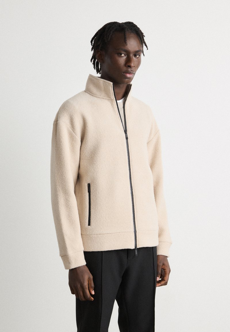 Beige fleece zip-up jacket with high collar, black zipper, and side pockets; relaxed fit with ribbed cuffs. Worn with dark trousers.