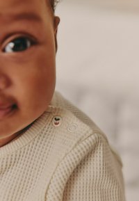 Šviesiai smėlinis, tekstūruotas mezginys su apvaliu iškirpimu. Yra dekoratyvinis mygtukas su žaliuias augalais ir baltu fono.