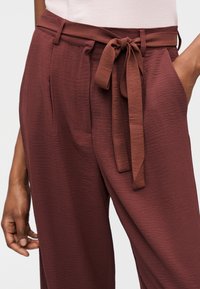 Γυναίκα που φοράει μακριά παντελόνια μπορντό με υφή και ζωνάρι από ύφασμα δεμένο στη μέση, με τα χέρια της να ακουμπούν κοντά στις τσέπες.