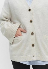 Krämfärgad kofta med v-ringning, fem bruna knappar, ribbade ärmslut och två framfickor. Mjuk textur, något oversized passform.