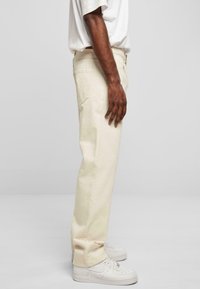 Pantalons beige à jambes droites en coton avec poches latérales. Associés à des baskets blanches, présentés de profil. Design minimaliste.