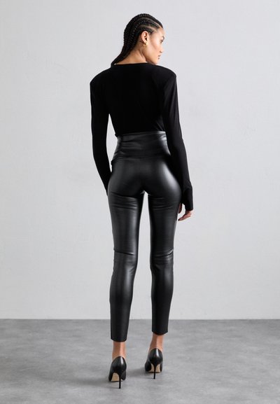 Sort langærmet top parret med højttaljede sorte kunstlæder leggings, der har en glat tekstur og tætsiddende design, accentueret med høje hæle.
